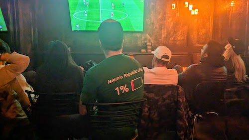 A group of friends, some in protest shirts, watching Iran play England on television