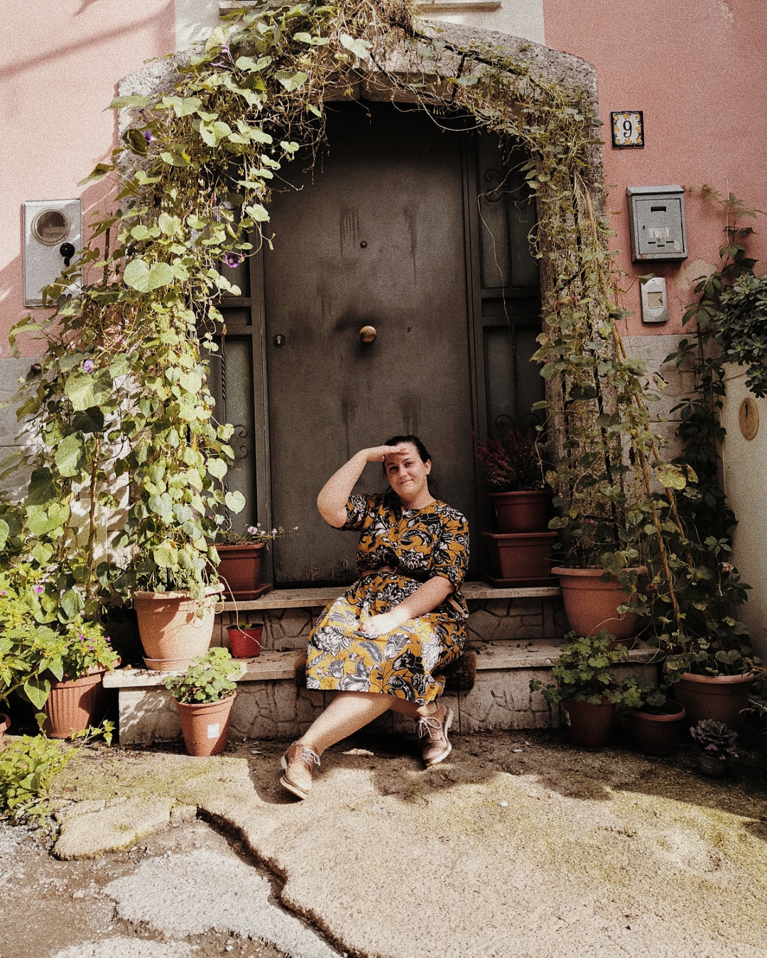 Ilaria è seduta fuori da casa sua, circondata da piante in vaso. Veste con un abito floreale color senape e scarpe marroni. I capelli sono raccolti in una coda e con una mano si scherma dal sole