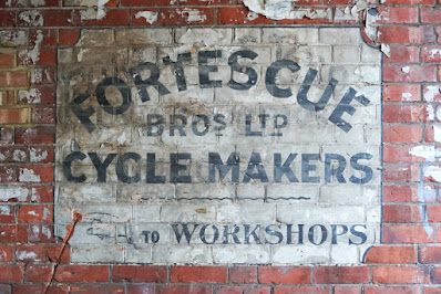 A ghost sign painted on red brick: a white background, with black text reading 'FORTESCUE BROS LTD CYCLE MAKERS. A manicule points left, with the text 'TO WORKSHOPS'