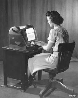 A black and white photograph of a woman seated at a desk and typing. 