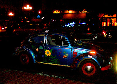 This picture is a side view of a classic blue Volkswagen Beetle the wheels are painted red.  Two flowers are also present.  Behind the Beetle can be seen the neon cityscape in the window of the McMillan Realty window.