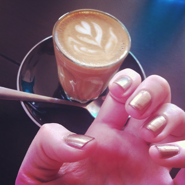 Photo of a piccolo coffee and my hand showing off my light bronze nail polish.