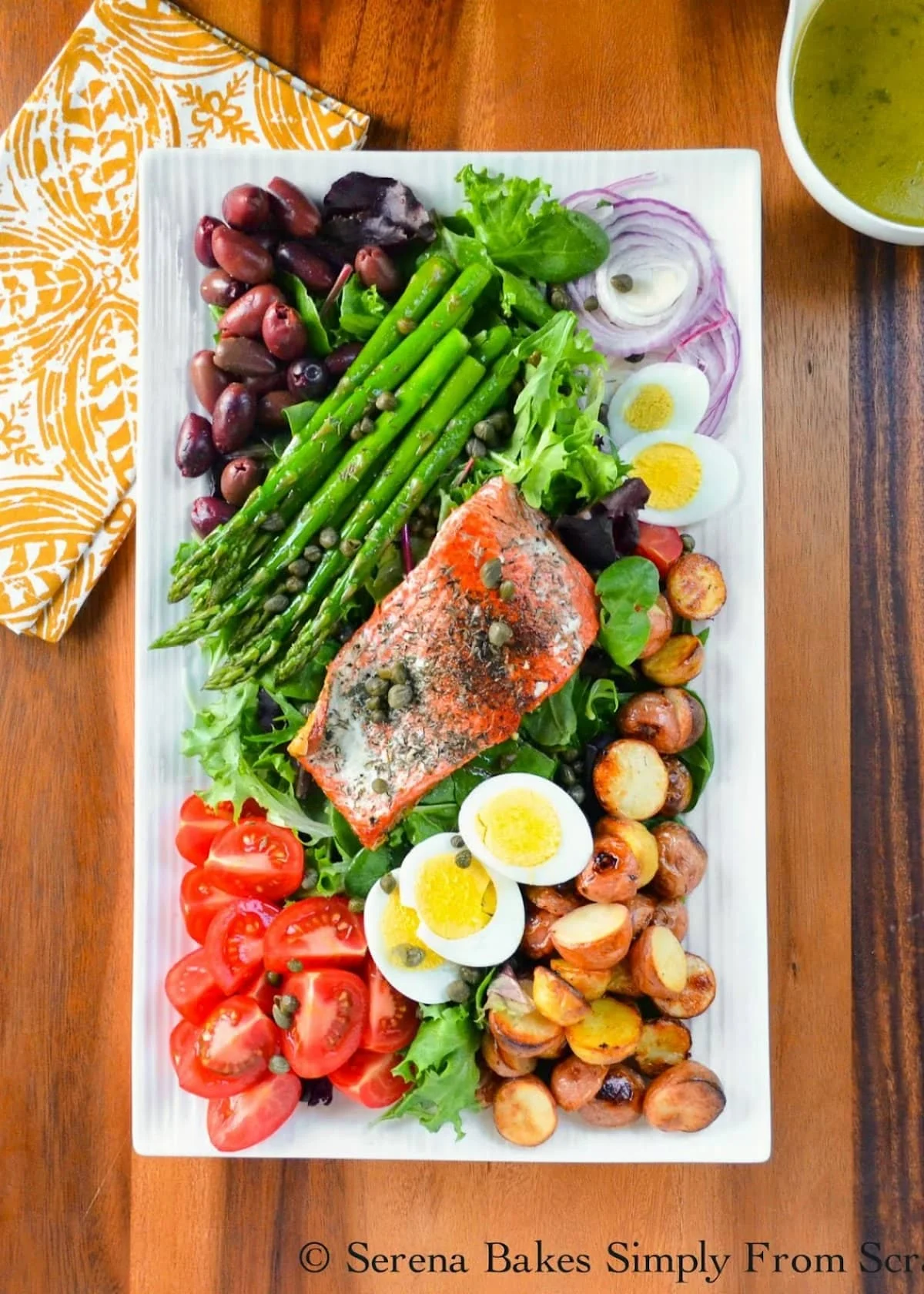 A photo looking down of Salmon Nicoise Salad on a white serving tray on a large wooden board with a white dish holding White Balsamic Vinaigrette in the top right corner of the photo.