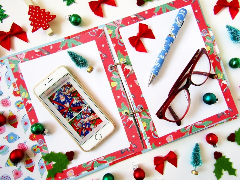 A Christmas flatlay image taken from above showing an open Christmas organiser layered with an  iPhone, pen, and a pair of red glasses, surrounded by small Christmas decorations like mini baubles, bows, and Christmas trees in red and green.