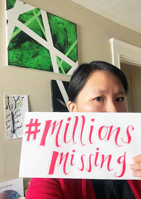 A woman holds a handmade sign that says "#Millions Missing" in red ink. The lower part of her face is partially covered by the sign, and the part you can see is half in light, half in shadow.