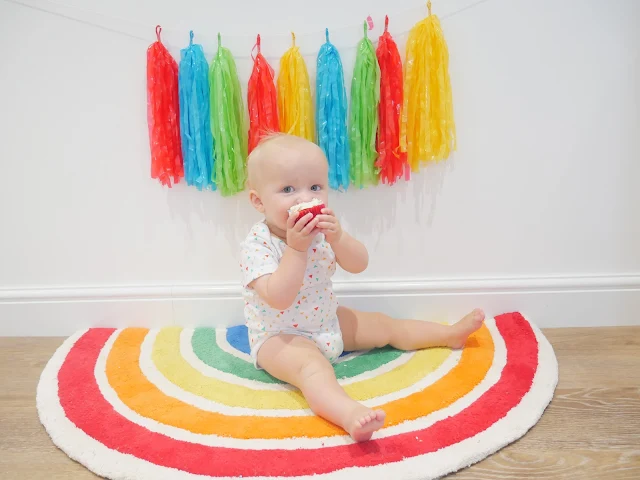 A rainbow rug sat on by a baby eating a cupcake