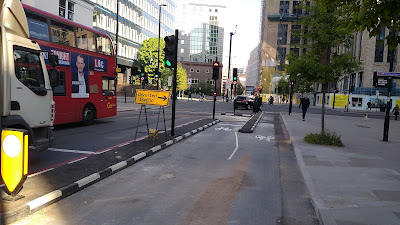 The two way cycle lane meets a junction. The road is on the right and bolt down islands protect cycle traffic. There are various traffic signals ahead. Traffic is moving through the junction.