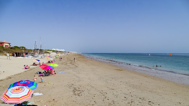 Playa llana de arena con gente con sombrillas y las azules aguas del mar a su frente.