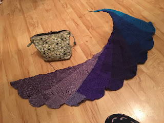 Tunisian crochet shawl spread out on the floor for a photo. The shawl is being worked in a gradient yarn in shades of blue, ranging from turquoise to navy, and purples, ranging from deep purple to lilac. The shawl is worked in long narrow wedge shaped panels ended with fan-shapes. The panels are offset, joined while working, giving the shawl an asymmetrical shape with the wider ends and fans forming the left side, and the narrow ends forming the right.