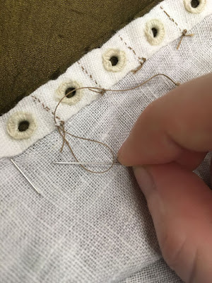 A close-up of a line of neat white eyelets along a double-folded hem in white linen, on top of green-gold silk. Lines of tiny tan stitches hold down the fabric between each pair of eyelets, and a white hand is using a needle to tie off the tail of the leftmost line of stitching at the inner fold of the hem.