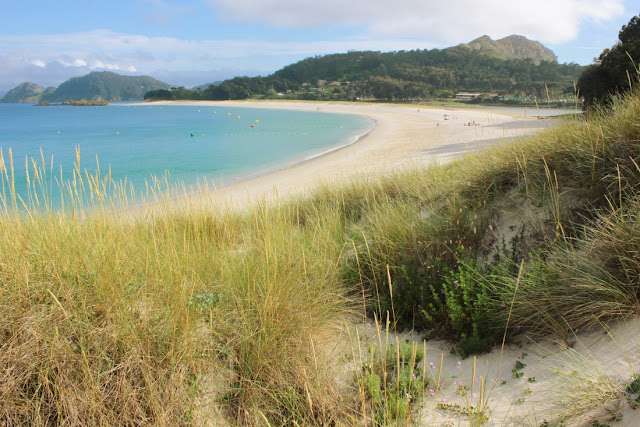 Widok na plażę Rodos, zajmującą łukowato wygiętą mierzeję, która łączy dwie z wysp Cies. Nad plażą wznosi się wzgórze z latarnią. W tle widać górzystą sylwetkę trzeciej z wysepek. 