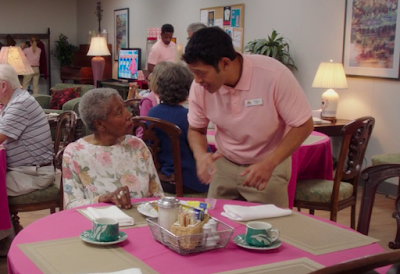 Pillboi, wearing a pink polo shirt and khakis, is talking to an old black woman with short gray hair, sitting at a table with a pink tablecloth and various food-related objects, like salt and pepper shakers, on it. They're at an old folks home and there are multiple people in the background.
