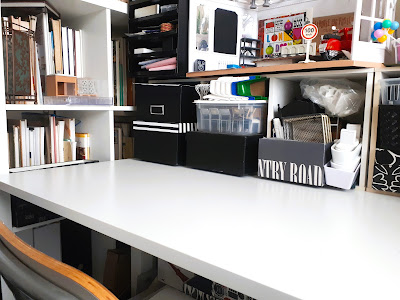 Clear work table with a bank of storage cubbies at the back filled with boxes and dolls house miniatures, and a roombox on top.