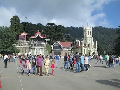 Visitors walking around The Ridge in Shimla, with the historic Christ Church, Shimla State Library, and a scenic hill backdrop under a bright blue sky.
