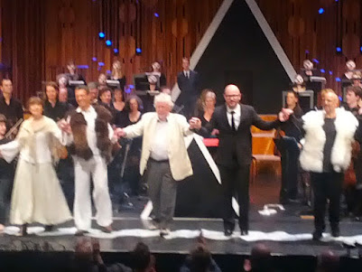 Blurry photo of a stage with a group of people taking bows, including baritone Roderick Williams, composer Harrison Birtwistle, and conductor Baldur Brönnimann.