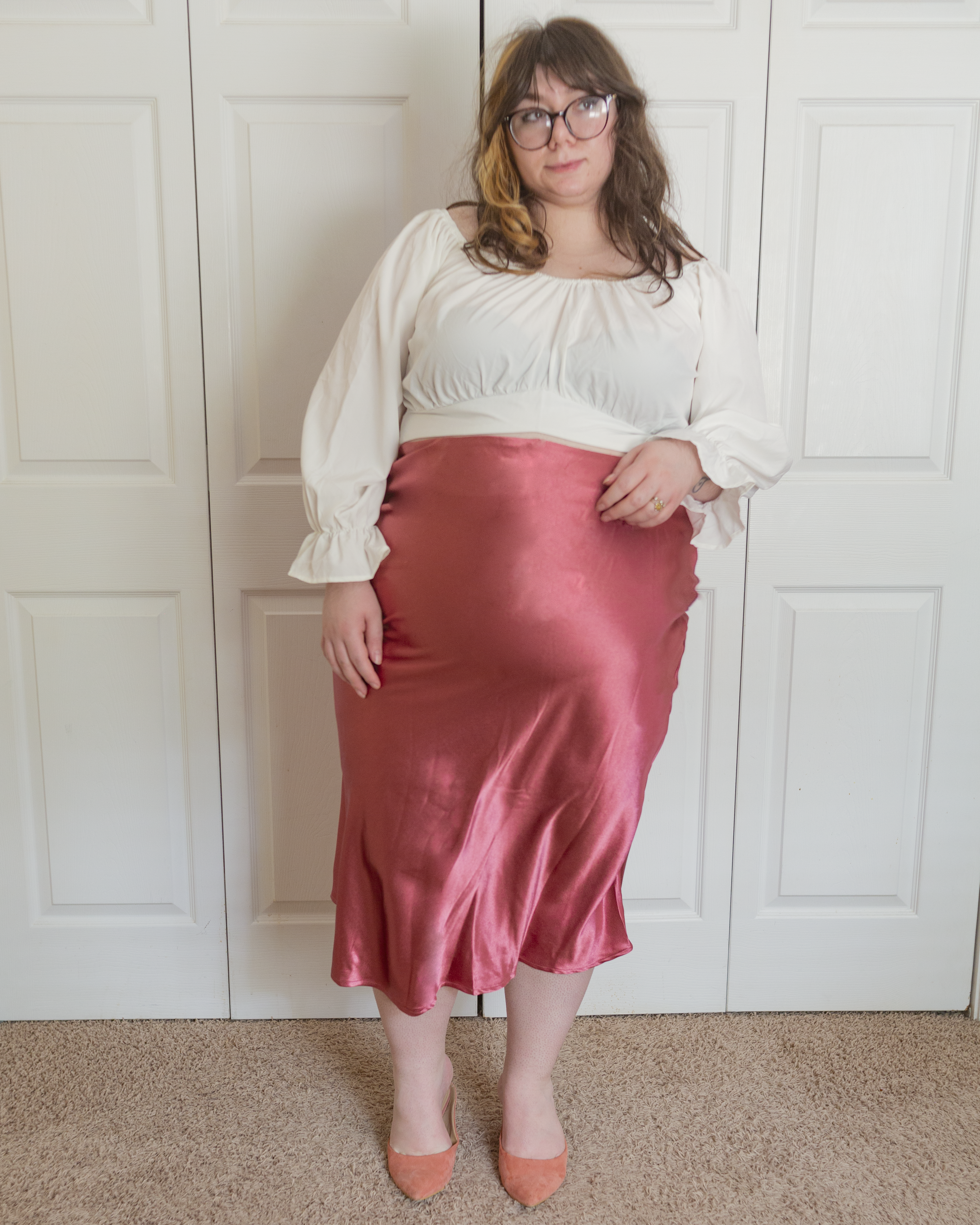 An outfit consisting of a white off the shoulder long bishop sleevbe blouse, a rosey pink satin slip skirt and pastel pink heeled clogs.