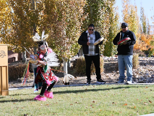 A young native american dancer in colorful attire dancing, with men drumming in the background