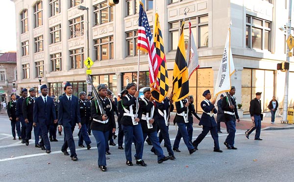 people in military uniforms--seemingly all Black and all male--march along a street in formation, carrying an American flag and various other flags, one bearing the name of a high school