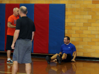 A man sitting on the gym floor in a blue shirt with a man in orange shirt talking to him