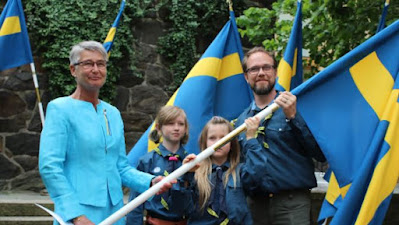 En scoutklädd man och två scoutklädda barn tar emot fana från landshövdingen