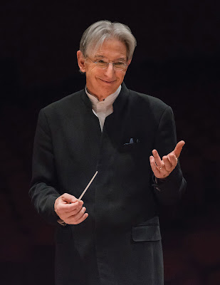 Photo of Michael TIlson Thomas. He is conducting, with a baton in his right hand, making a gesture with his left. He is a gray-haired, slender man with a smile on his face.