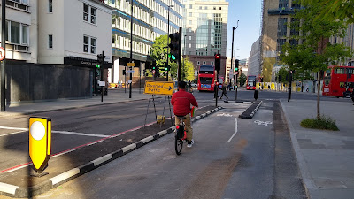 The two way cycle lane meets a junction. The road is on the right and bolt down islands protect cycle traffic. There are various traffic signals ahead. A cyclist can be seen going ahead.