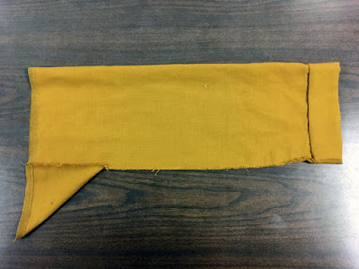 A photo from above of a perfectly rectangular mustard-brown sleeve with a small right-triangle gusset at lower left and a deep double-folded cuff at right.