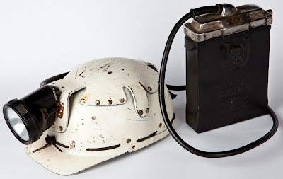 A white peaked safety helmet, clearly well used, with a safety lamp and battery attached