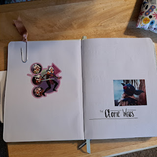A cover page in a blank book with a sticker of the Star Wars character, Ahsoka, and a picture of the Sesame Street character, Grover with Yoda ears.