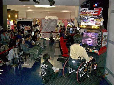 Image of a Namco/Wangan Midnight Maximum Tune 3 arcade racing game - with the seat removed and replaced with a wheelchair ramp. It seems the foot pedals have been replaced by a hand control.