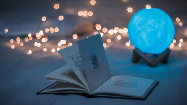 A hardcover book is open on a white blanket. In the background is a blue globe and white christmas lights.