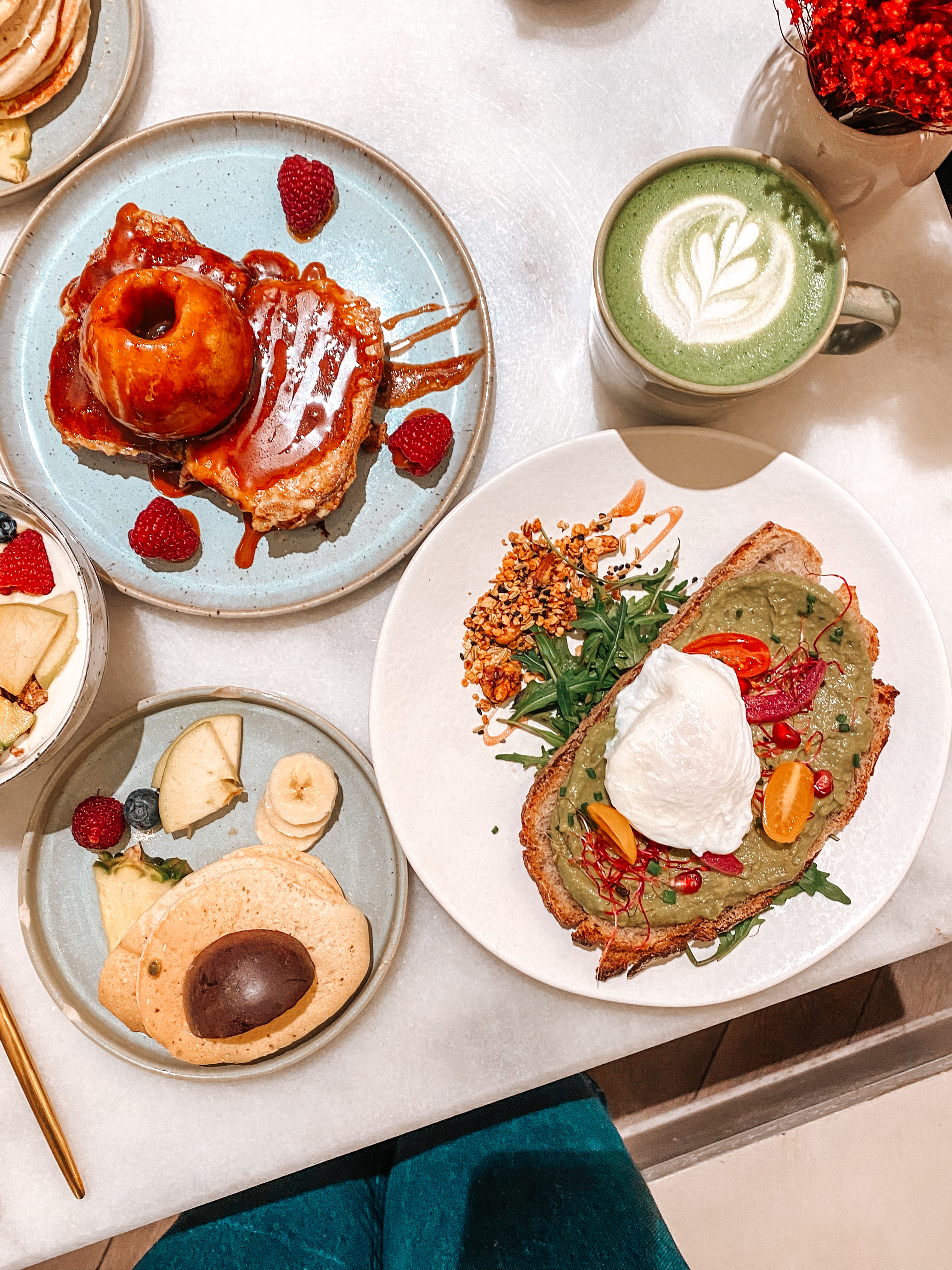 en bas à droite un avocado toast avec un oeuf poché et des tomate sur le dessus avec un matcha latte dans une tasse verte et à droite des pancakes avec une pomme caramélisée sur le dessus