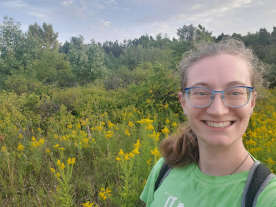 A picture of me, a white young woman with blonde hair in a ponytail and glasses, in front of a field of goldenrod with trees in the background