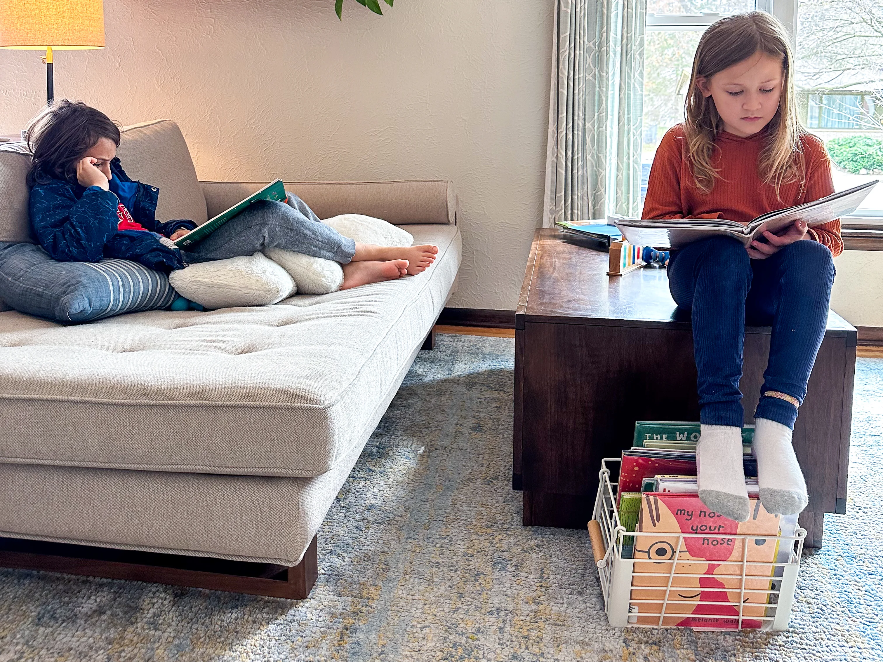 Two Montessori elementary kids sit and read picture books in the living room of their Montessori home