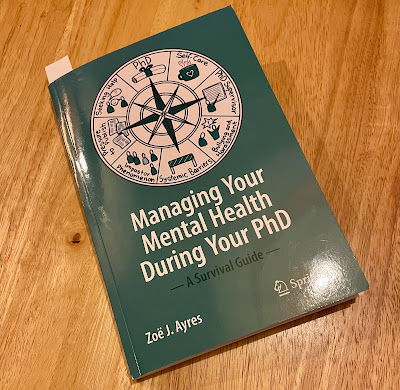 Copy of the book Managing your Mental Health During Your PhD - A Survival Guide by Dr Zoë J Ayres, placed at an angle on a wooden surface.