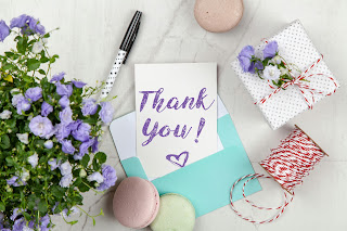 Thank you note on table with pen next to a wrapped gift, cookies and flowers