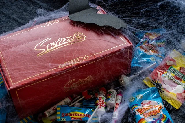 A red Swizzels box with gold writing surrounded by sweets. All of it is covered in spiders webs, plastic spiders and a cut out bat