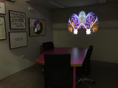 An office space with art on the walls has a projection of animal skulls and flowers in an oval shape on one wall, above a pink table.