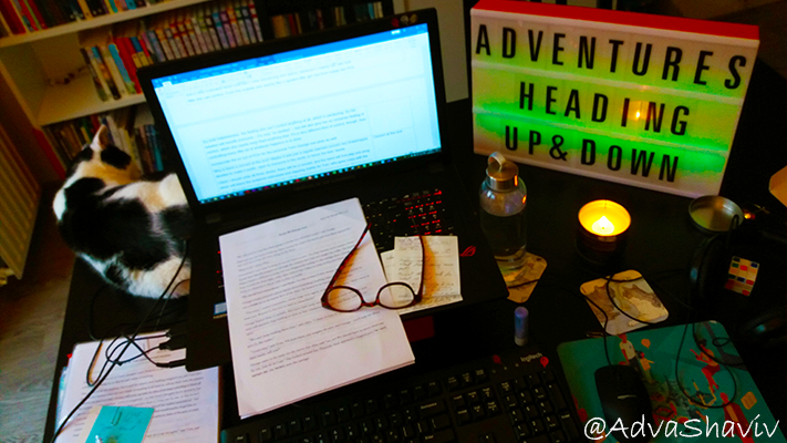 A desk with an open laptop, papers, cat, reading glasses, lightbox, candle, water bottle.