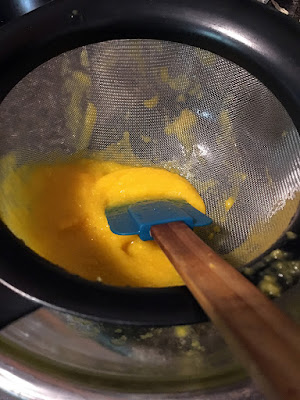 A black-handled strainer with about half a cup of thick mango puree in the bottom, and a blue silicone spatula resting on the bottom of the strainer.