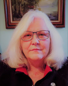 White woman with shoulder-length white hair and glasses, wearing a black top with a red collar. A framed painting is partially visible behind her.