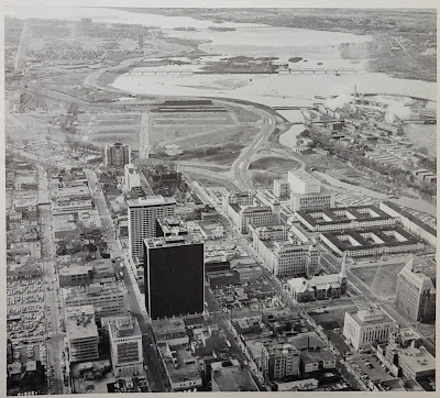 Aerial view from above looking west in 1967. Centretown/Uppertown in the foreground including Place De Ville and Library and Archives Canada; LeBreton Flats in the midground; Ottawa River, Lemieux Island and Mechanicsville in the distance. Chaudière Island industry at work. West of Bay, Wellington Street becomes the Ottawa River Parkway with an offramp connection to Wellington Street through LeBreton Flats.