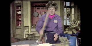 Julia Child is bashing some dough on a counter, with a rolling pin