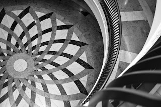 A black and white photograph of a marble floor from the point of view of a second floor interior balcony. The balconies and floor beneath are round, the pattern on the floor an overlapping floral geometric pattern in three shades of marble. The balconies have iron railings and the middle of the floor design is off-centre in the image.