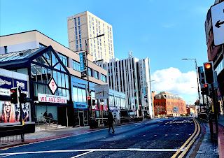 A photo of some light brown square shops with a square pyramid based silver glass and metal store with m in white font in a red circle with a grey road with white stripes down the street on a bright background