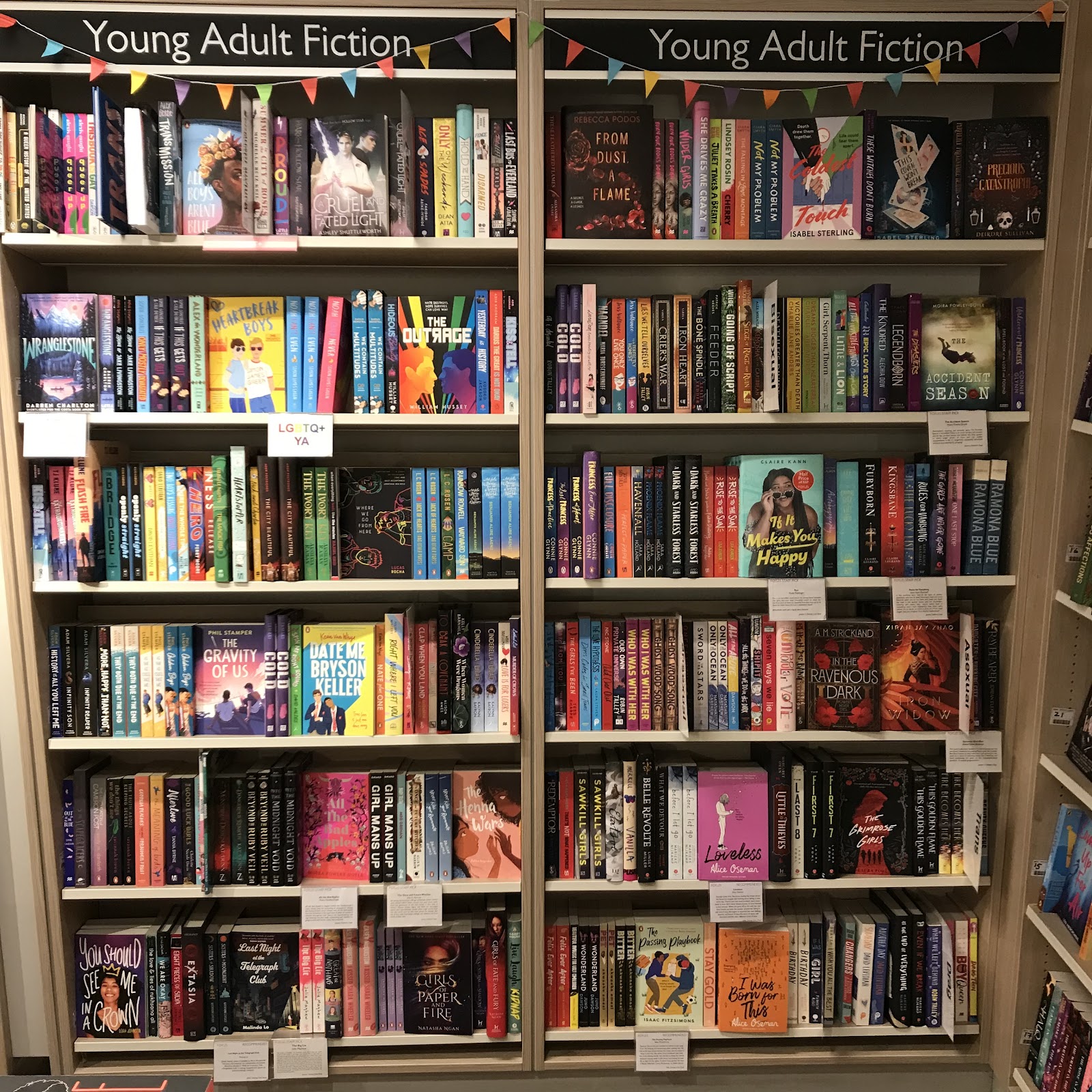 The LGBTQ+ YA section at Foyles, Charing Cross Road, face on. It consists of two bays/bookcases. The first bay on the left has LGBTQ+ teen non-fiction, multiple identities, gay, and lesbian. The second bay has the last lesbian books, bisexual, pansexual, asexual, trans, non-binary and other genders, and drag. There are various staff pick shelf talkers on the shelves recommending books.