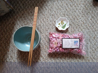 A bowl with chopsticks, a little dish with a fern and flowers on it and a plastic bag filled with pink potpourri.