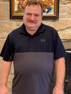 Man with brown hair and mustache wearing a gray and black short-sleeved shirt stands in front of a stone wall