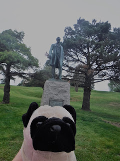 a weathered green statue of Abraham Lincoln stands atop a stone pedestal in a grassy park between two trees behind a plush pug.