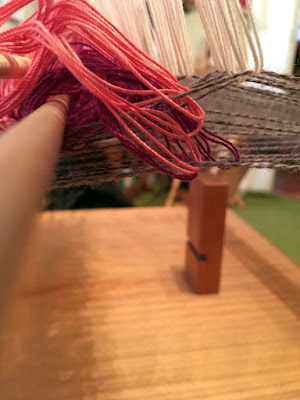 A view through the shallow, tangled triangular tunnel formed by raised and flat grey warp threads. The raised threads are held up by white loops of fine smooth string, barely visible over the piles of saturated fuchsia and salmon-pink loops sitting on and through the threads forming the left side of the triangle. 